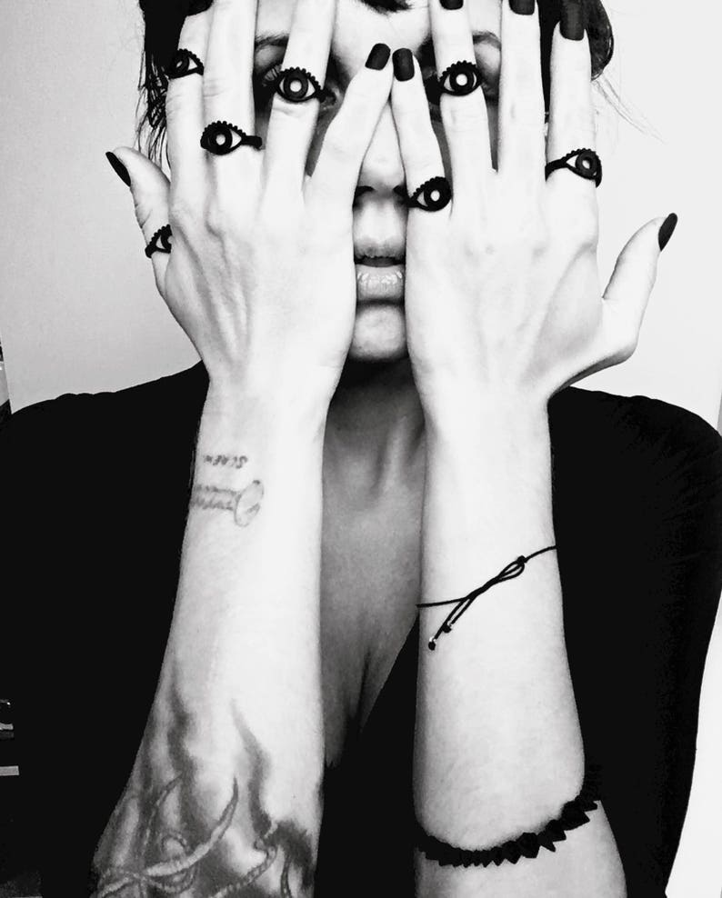 Matte black 3d printed eye ring displayed on a tattooed model's hands with long black nails covering her face