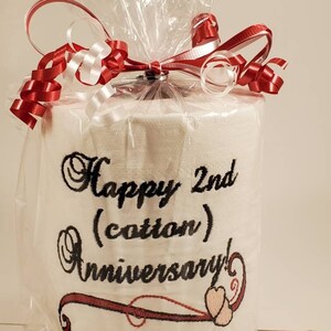 May include: A white toilet paper roll wrapped in clear plastic with a red and white ribbon. The toilet paper has black embroidery that says "Happy 2nd (cotton) Anniversary!" with a red and pink heart design.