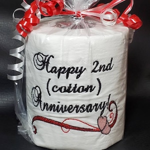 May include: A white toilet paper roll with red and white ribbon wrapped around it. The toilet paper has black embroidery that says "Happy 2nd (cotton) Anniversary!" with a red swirl and two red hearts.