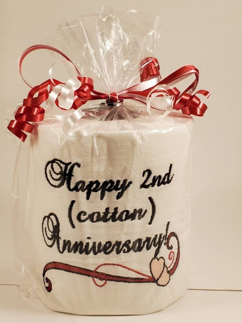 May include: A white toilet paper roll wrapped in clear plastic with a red and white ribbon. The toilet paper has black embroidery that says "Happy 2nd (cotton) Anniversary!" with a red and pink heart design.