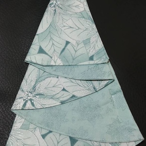 May include: A folded fabric Christmas tree in shades of teal and white. The tree is made from a fabric with a floral pattern, with layers of fabric creating the tree shape. The background is black.
