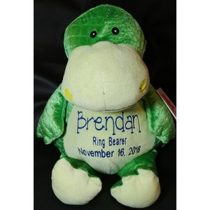 May include: A green plush dinosaur toy with a cream-colored belly. The name "Brendan", the text "Ring Bearer", and the date "November 16, 2018" are embroidered in blue on the belly. The dinosaur has black eyes and yellow accents.