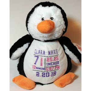 May include: A plush penguin toy with black and white fur, an orange beak and feet. The penguin has the name "Clara Marie" embroidered in purple, along with weight, length, time, and date details.