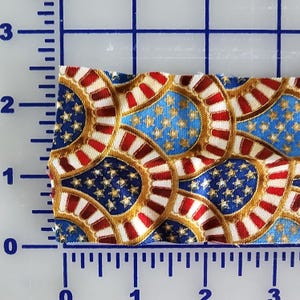 May include: Close-up of a fabric swatch featuring a patriotic design. The pattern includes blue fields with gold stars, red and white stripes, and gold borders. The fabric is measured with an inch ruler.