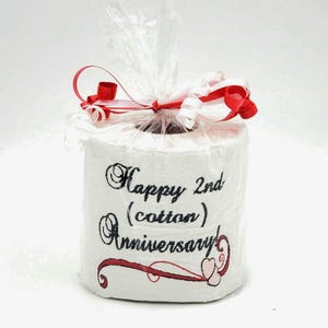 May include: A white toilet paper roll with a red ribbon and a black embroidered message that says "Happy 2nd (cotton) Anniversary" with two pink hearts.