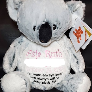 May include: A plush koala bear in shades of gray and white, with a black nose and embroidered details. The bear has the name "Asia Ruth" and the date "October 19 2020" embroidered in pink and black thread, along with a quote from Jeremiah 1:5.