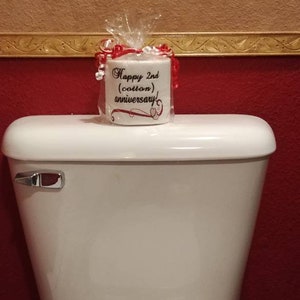 May include: A white toilet paper roll with a red ribbon and a white label that says "Happy 2nd (cotton) anniversary!" The toilet paper roll is sitting on top of a white toilet tank.