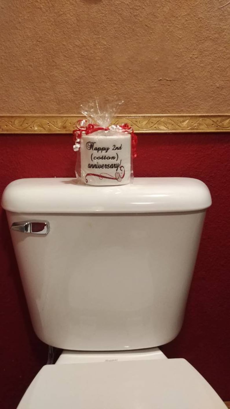 May include: A white toilet paper roll with a red ribbon and a white label that says "Happy 2nd (cotton) anniversary!" The toilet paper roll is sitting on top of a white toilet tank.