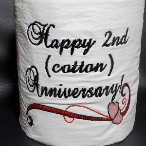 May include: A white toilet paper roll with the text "Happy 2nd (cotton) Anniversary!" embroidered in black thread. A red and black swirl design with a pink heart is embroidered below the text.