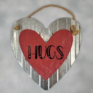 Puede incluir: Decoración de pared en forma de corazón con un fondo de metal corrugado y un corazón de madera rojo superpuesto. La palabra "HUGS" está impresa en escritura negra. Se adjunta una cuerda de yute para colgar.