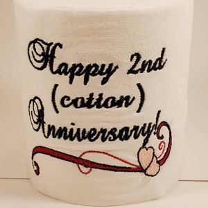May include: A white toilet paper roll with black embroidery that says "Happy 2nd (cotton) Anniversary!" with a red and pink swirl design.