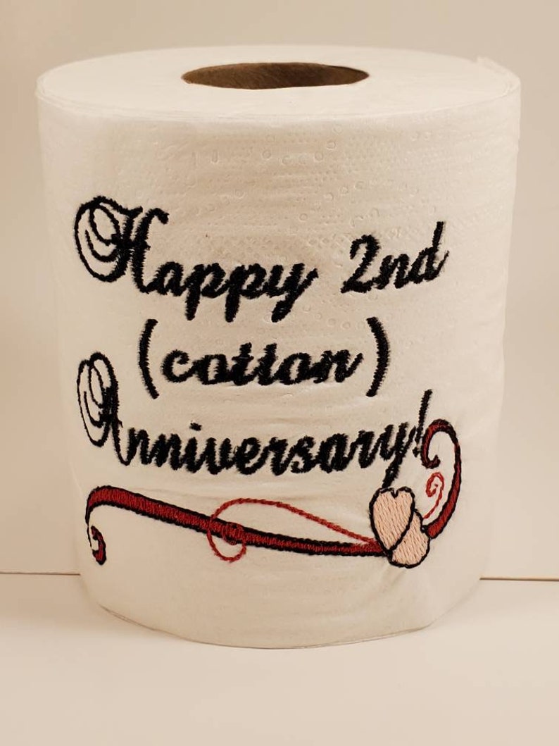 May include: A white toilet paper roll with black embroidery that says "Happy 2nd (cotton) Anniversary!" with a red and pink swirl design.