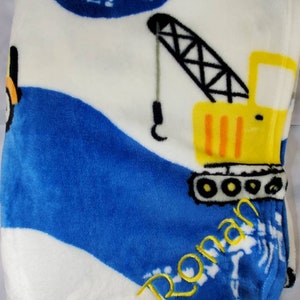 Puede incluir: Una manta suave y afelpada con un tema de construcción. La manta tiene un fondo blanco con olas azules y una grúa amarilla. El nombre "Ronan" está bordado en escritura amarilla. La manta está diseñada para la comodidad y el calor.