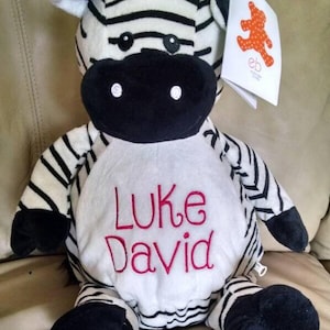 May include: A plush zebra stuffed animal with black and white stripes, a black nose, and black feet. The zebra has the name "Luke David" embroidered in red on its white belly. A tag with a red figure is attached.