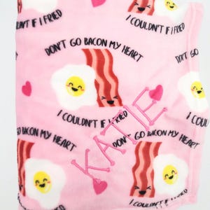 May include: Pink fleece blanket with a repeating pattern of cartoon bacon and eggs with the text "Don't go bacon my heart" and "I couldn't if I tried". The blanket is personalized with the name "Katie" in pink thread.