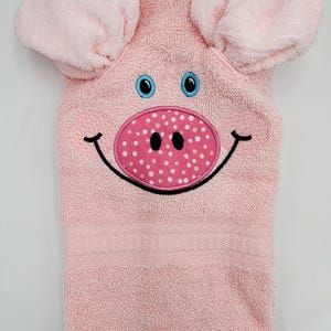 May include: A pink hooded towel with a pig face design. The towel features large pink ears, blue eyes, a pink and white polka-dotted snout, and a smiling mouth. The towel is made of a soft, absorbent material.