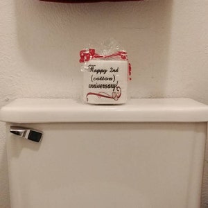 May include: A white toilet paper roll with a red ribbon and the text "Happy 2nd (cotton) anniversary!" on a white toilet tank.