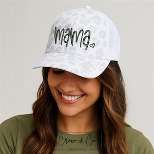 Puede incluir: Gorra de camionero blanca con un estampado de leopardo en el panel frontal y una parte trasera de malla blanca. La palabra "mama" está bordada en escritura verde oscuro con un pequeño corazón. La gorra tiene una visera curva y un botón en la parte superior.