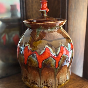 Könnte beinhalten: Ein glasierter Keramiktopf mit Deckel. Der Topf ist überwiegend braun mit auffälligen roten und weißen wellenartigen Mustern. Der Deckel und der Knauf sind leuchtend rot. Der Topf steht auf einer Holzoberfläche.