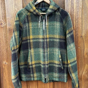 May include: A hooded jacket with a plaid pattern in shades of green, black, and yellow. The jacket has a full zipper, two front pockets, and white drawstrings. The jacket is hanging on a wooden hanger.