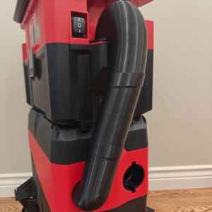 Könnte beinhalten: Ein rot-schwarzer Akku-Staubsauger mit einem auffälligen schwarzen Schlauchaufsatz. Der obere Teil ist mit FUEL BRUSHLESS und M18 beschriftet. Er verfügt über einen schwarzen Netzschalter an der Seite und steht auf einer Basis mit kleinen Rädern.