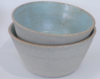 May include: Two ceramic bowls stacked on top of each other. The top bowl is light blue and the bottom bowl is a light gray with a speckled texture.
