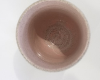 May include: A light pink ceramic cup with a speckled glaze. The cup is empty and the inside is visible.