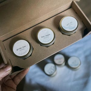 Puede incluir: Un juego de tres pequeños botes redondos plateados con etiquetas blancas dentro de una caja de cartón. Las etiquetas dicen "golden hour", "fern canyon" y "salt air". La caja está abierta, revelando los botes. El fondo es de un color suave y neutro.