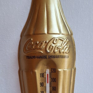 May include: Gold Coca-Cola bottle shaped thermometer with a red liquid inside. The thermometer reads from 120 degrees Fahrenheit down to 30 degrees Fahrenheit. The thermometer has the text "Coca-Cola" and "Trade-Mark Registered" on it.