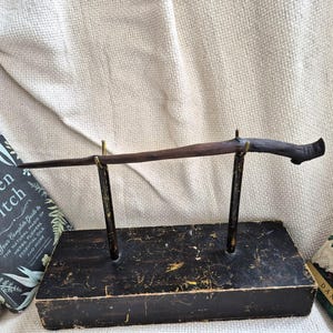 May include: A dark wooden wand displayed on a black stand with brass supports. The wand is presented with two books, one titled "The Green Witch" with botanical illustrations and text. The other book is partially visible.