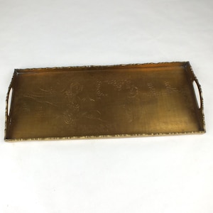 May include: A rectangular brass serving tray with a metallic gold finish, featuring integrated side handles and a textured, scalloped edge. The center of the tray displays a faint, etched design of a figure and floral motifs on its flat surface.