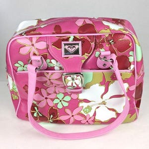 May include: A rectangular handbag featuring a vibrant pink base with a multi-colored floral pattern in shades of white, light green, and dark red. The bag includes two pink shoulder straps, a silver buckle closure, and a Roxy logo metal plaque.