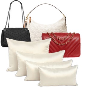 Puede incluir: Cuatro fundas de almohada de sat&eacute;n crema dispuestas en forma de pir&aacute;mide. En el fondo, un bolso de mano acolchado negro con una correa de cadena, junto con un bolso de mano estampado blanco y marr&oacute;n y un bolso de mano acolchado rojo con una correa de cadena dorada.