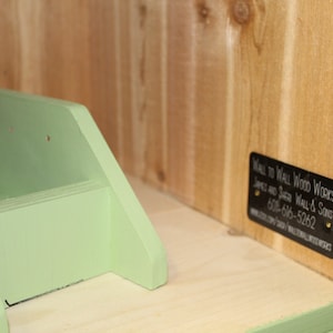 May include: A wooden shelf unit with a light green painted accent. The unit has a small shelf and a back panel with a black label that reads "Wall to Wall Wood Works". The unit is made of natural wood.