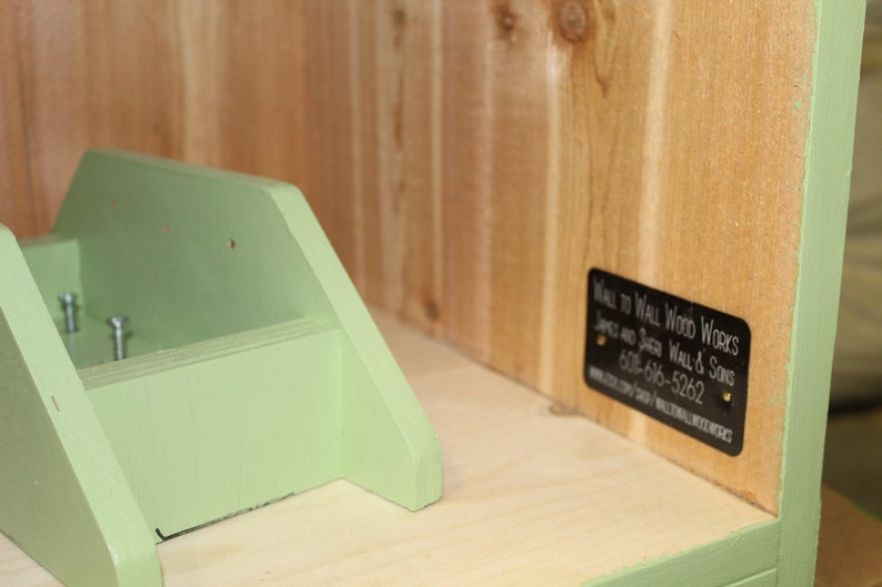May include: A wooden shelf unit with a light green painted accent. The unit has a small shelf and a back panel with a black label that reads "Wall to Wall Wood Works". The unit is made of natural wood.