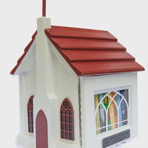 May include: A white wooden church-shaped Little Free Library with a red roof and cross. The library has a door and two arched windows with red frames. The front window displays books. The text "LittleChurchLibrary.org" is visible.