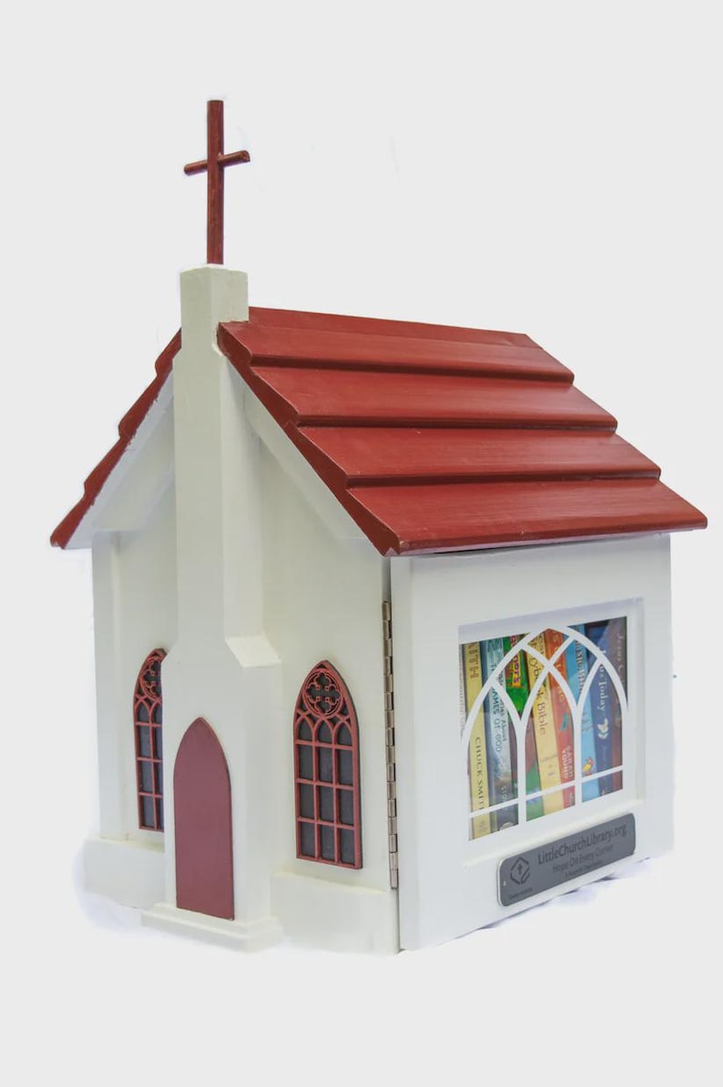 May include: A white wooden church-shaped Little Free Library with a red roof and cross. The library has a door and two arched windows with red frames. The front window displays books. The text "LittleChurchLibrary.org" is visible.