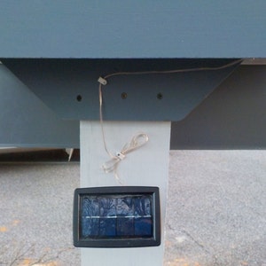 May include: A close-up of a solar powered light fixture mounted on a white post. The light fixture has a black rectangular solar panel and a white wire running from the panel to the top of the post.