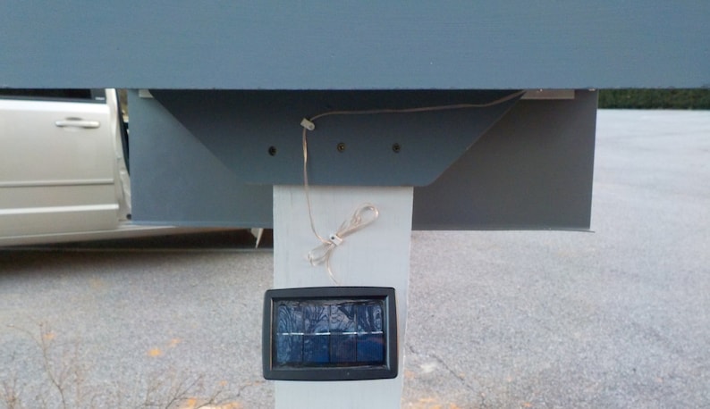 May include: A close-up of a solar powered light fixture mounted on a white post. The light fixture has a black rectangular solar panel and a white wire running from the panel to the top of the post.
