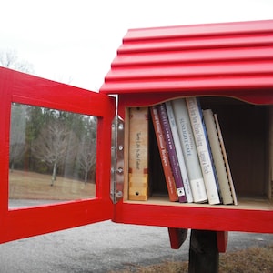 May include: A red wooden free library with a hinged door and a glass window. The library is filled with books with titles like "Sunlight Cafe" and "A Taste of Food".