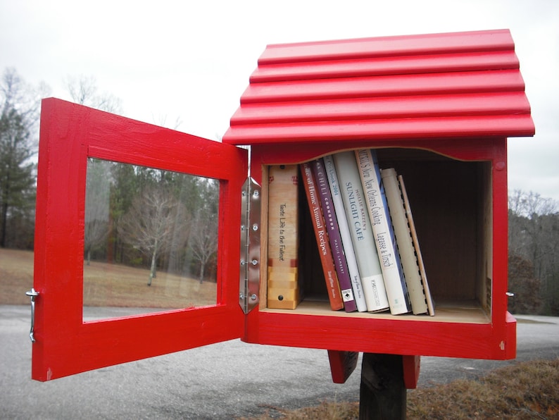 May include: A red wooden free library with a hinged door and a glass window. The library is filled with books with titles like "Sunlight Cafe" and "A Taste of Food".