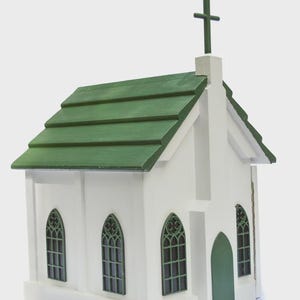 May include: A white wooden church model with a green roof and a cross on top. The church has arched windows with green trim and a green door. The model is a miniature replica of a church building.