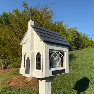 May include: A white, miniature church-shaped structure with a dark gray roof and a cross on top. It features arched windows and a small door. The structure is mounted on a white post and set in a grassy area.