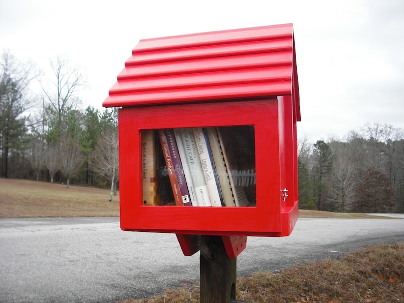 May include: A red wooden book exchange box with a pitched roof and a glass front. The box is filled with books with titles like "Night Cafe" and "Sweet Louisiana Food".