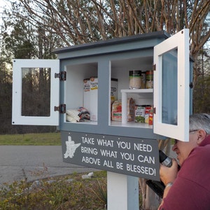 May include: A gray and white wooden box with two doors, one open, revealing shelves stocked with canned goods. The box has a sign that reads "TAKE WHAT YOU NEED BRING WHAT YOU CAN ABOVE ALL BE BLESSED!"