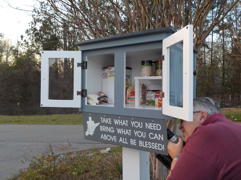 May include: A gray and white wooden box with two doors, one open, revealing shelves stocked with canned goods. The box has a sign that reads "TAKE WHAT YOU NEED BRING WHAT YOU CAN ABOVE ALL BE BLESSED!"