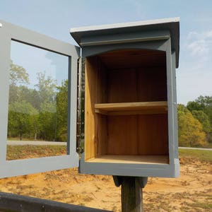 May include: A small, gray wooden Little Free Library with a clear glass door, open to reveal a shelf inside. The library is mounted on a wooden post and set against a backdrop of trees and a sunny sky.