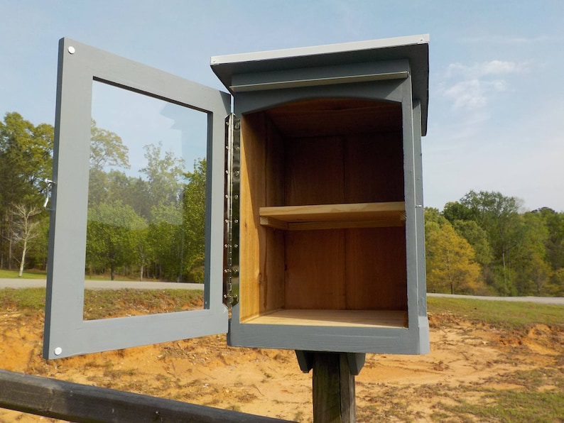 May include: A small, gray wooden Little Free Library with a clear glass door, open to reveal a shelf inside. The library is mounted on a wooden post and set against a backdrop of trees and a sunny sky.