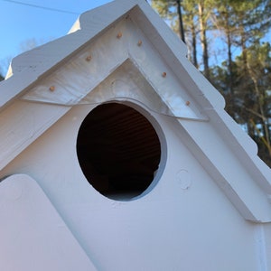 May include: White birdhouse with a round entrance hole and a decorative triangular roof. The roof has a white painted surface with small gold accents.