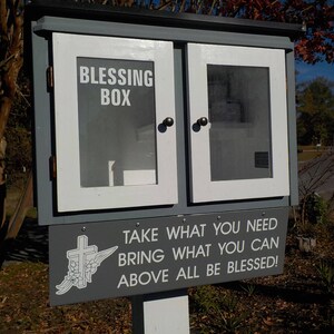 May include: A wooden box with a white door and a sign that reads "Blessing Box Take What You Need Bring What You Can Above All Be Blessed!"